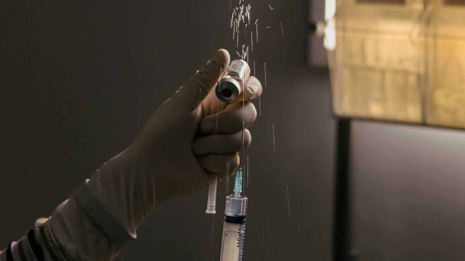 A gloved scientist in a dimly lit laboratory uses a syringe to prepare a solution.